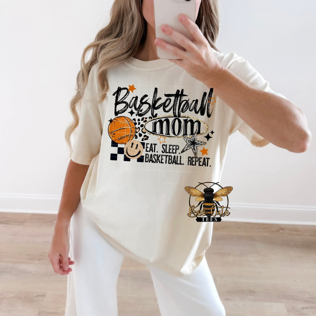 A woman takes a mirror selfie wearing a cream-colored t-shirt with a 'Basketball Mom' graphic. The graphic features a basketball,  a star, and the words 'EAT.