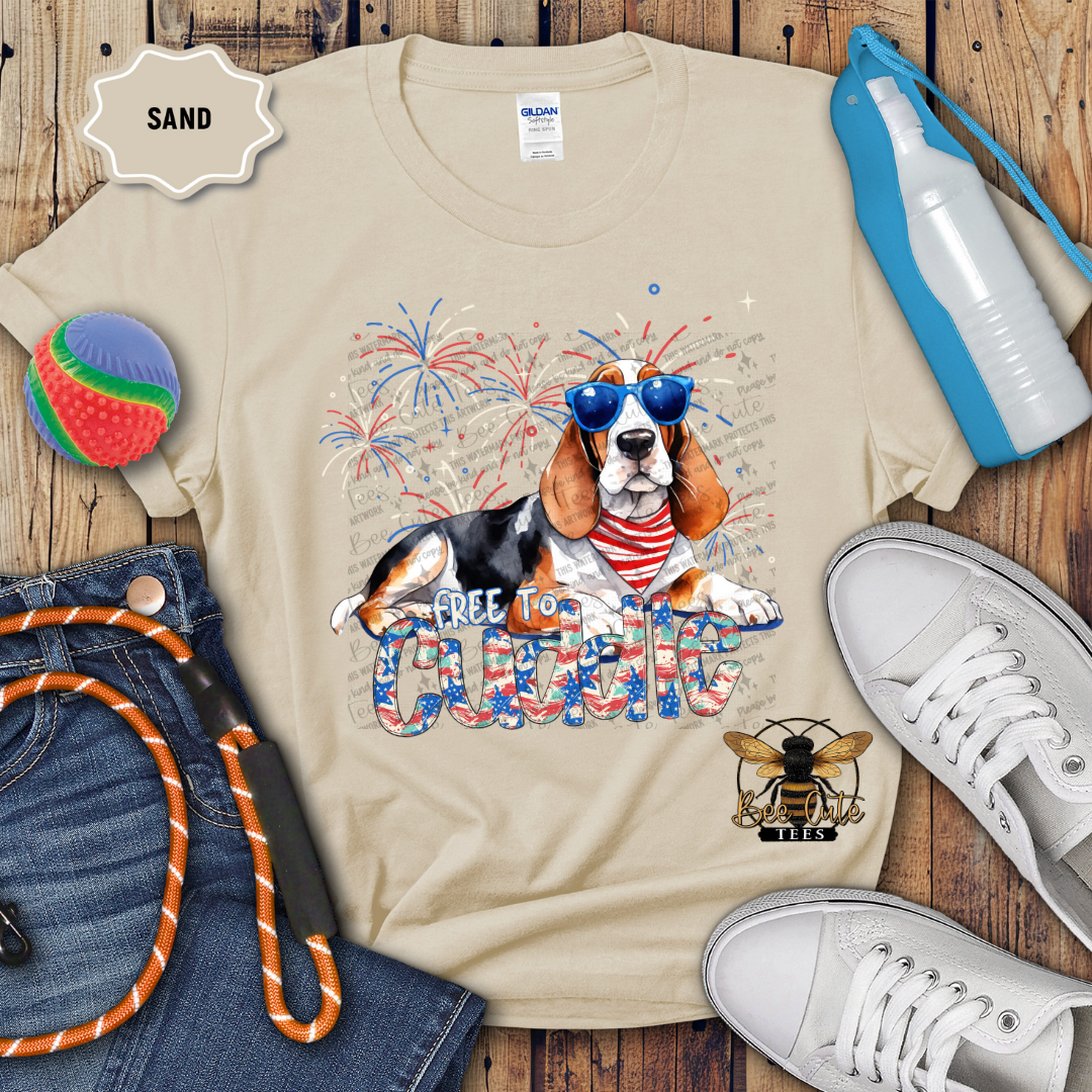 A beige colored t-shirt with a graphic print of a Bassett Hound wearing sunglasses and the text 'FREE TO Cuddle'. The shirt is displayed with a pair of jeans, a pair of white sneakers, a blue water bottle, a red and white toy, and a 'SAND' colored label.