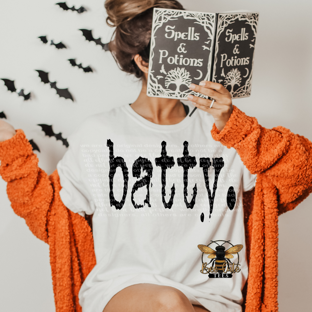 Person wearing a white shirt with 'batty' text, holding 'Spells & Potions' books, with Halloween decorations in the background.