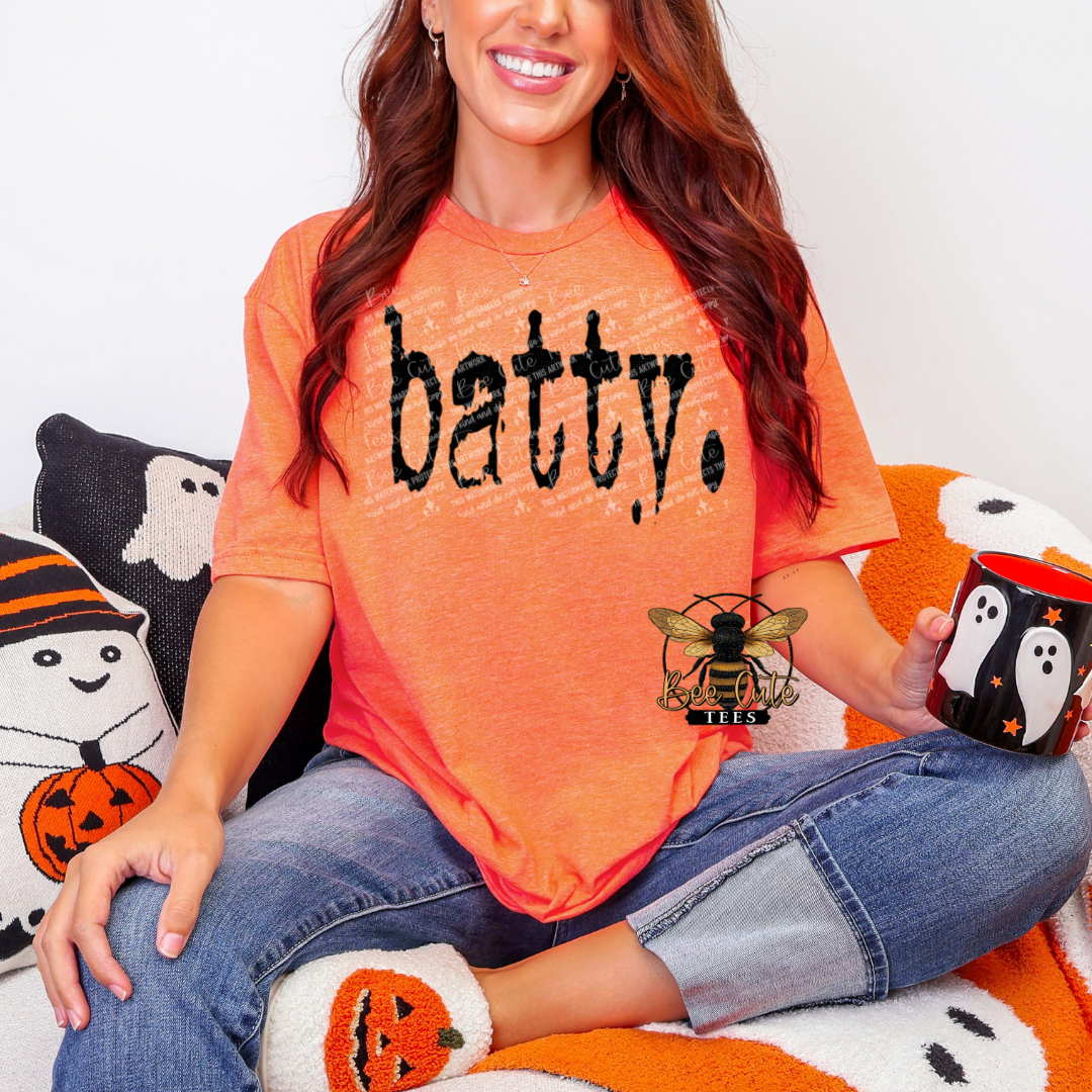 Woman wearing an orange 'batty.' shirt holding a Halloween-themed mug.