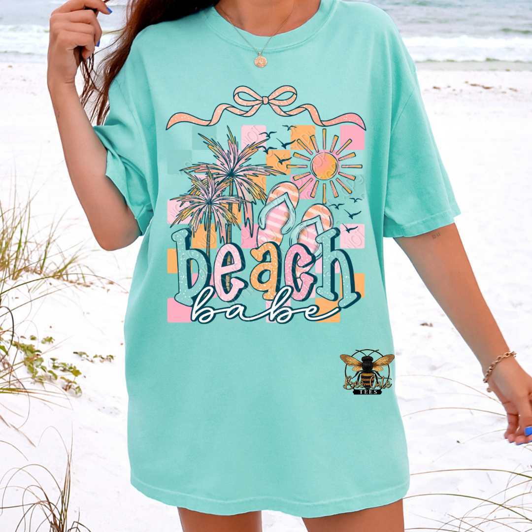 Person wearing a light green t-shirt with 'Beach Babe' design on a beach.