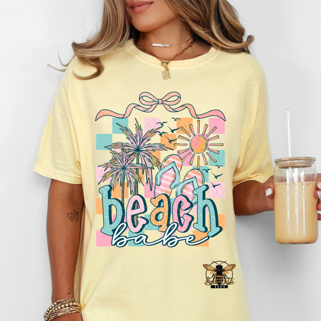 Person wearing a yellow t-shirt with 'beach babe' design, holding a drink.