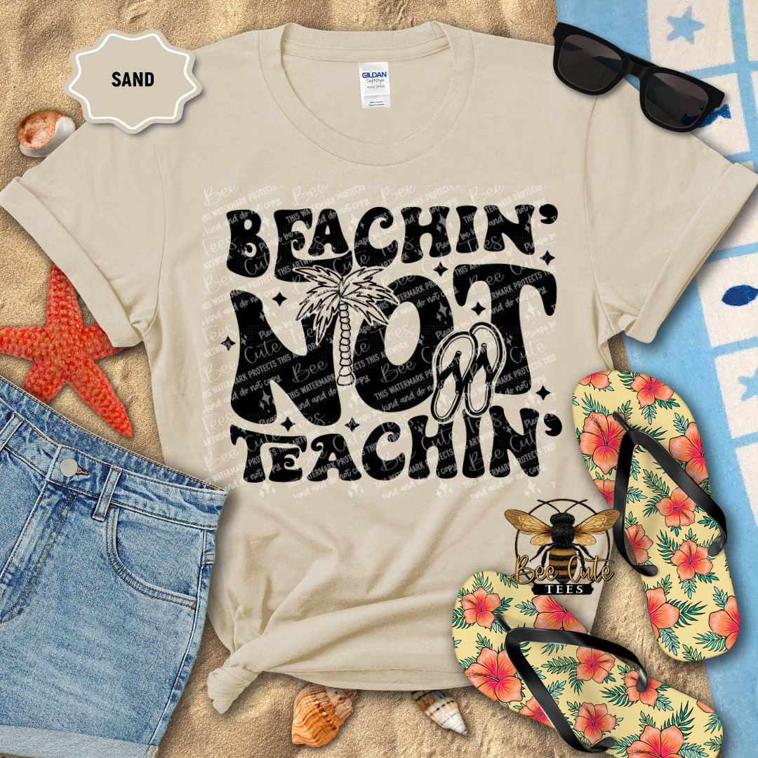 Beige t-shirt with 'Beachin' Not Teachin' design on sand with beach accessories.