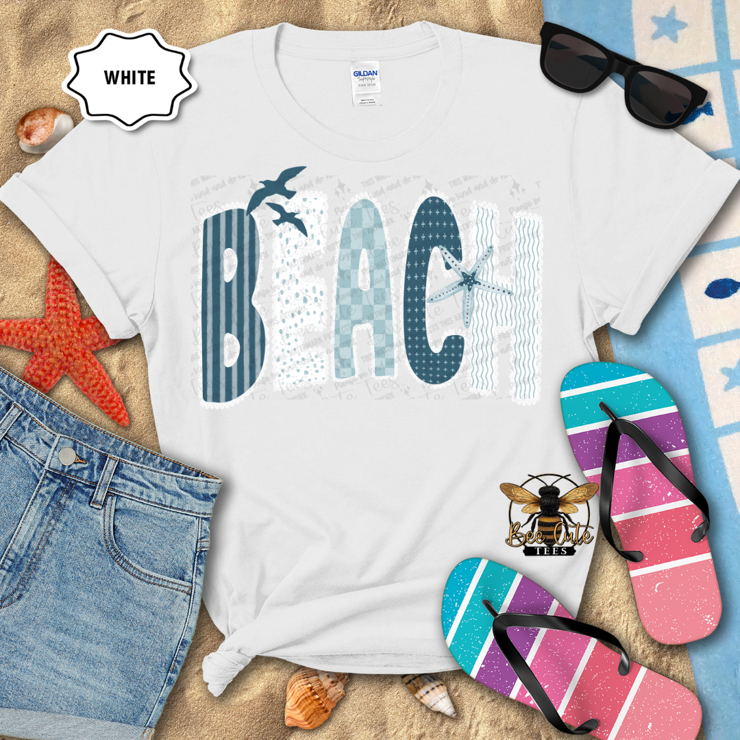 White t-shirt with 'BEACH' design on a sandy background with flip-flops and sunglasses.