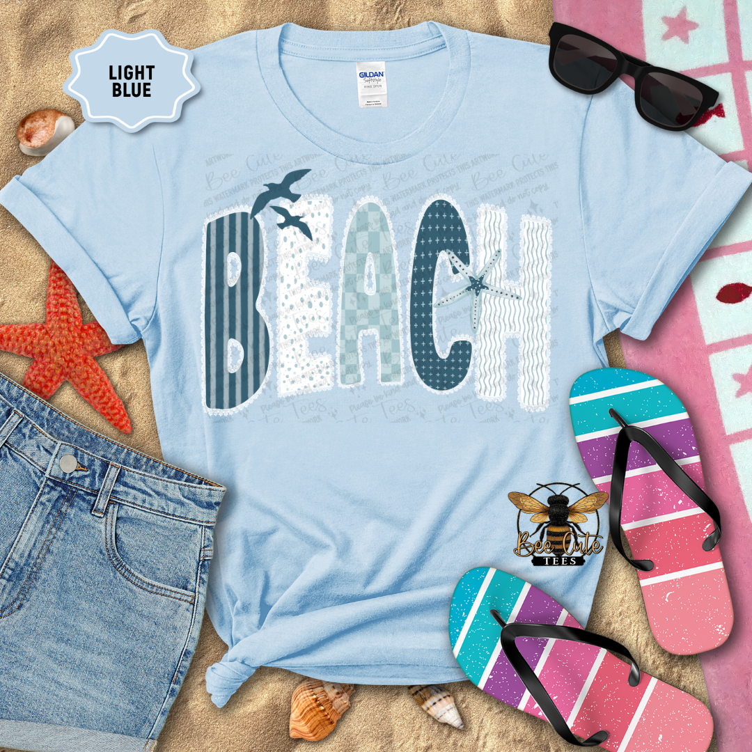Light blue t-shirt with 'BEACH' design on a sandy background with beach accessories.
