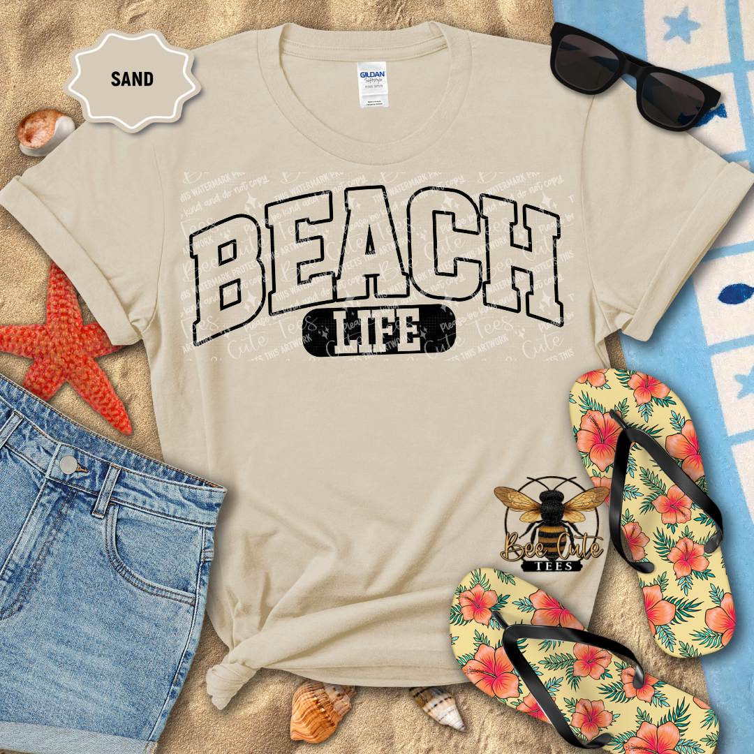 Beach-themed t-shirt with 'BEACH LIFE' text, surrounded by beach accessories on sand.