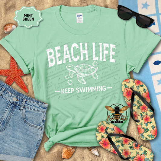 Mint green t-shirt with 'Beach Life Keep Swimming' design on a sandy background with flip-flops and sunglasses.