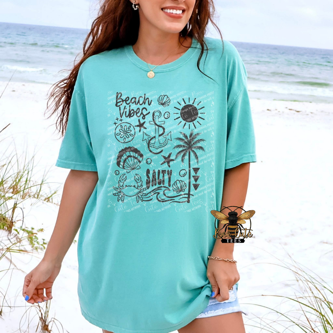 Woman wearing a turquoise t-shirt with beach-themed graphics on a beach.