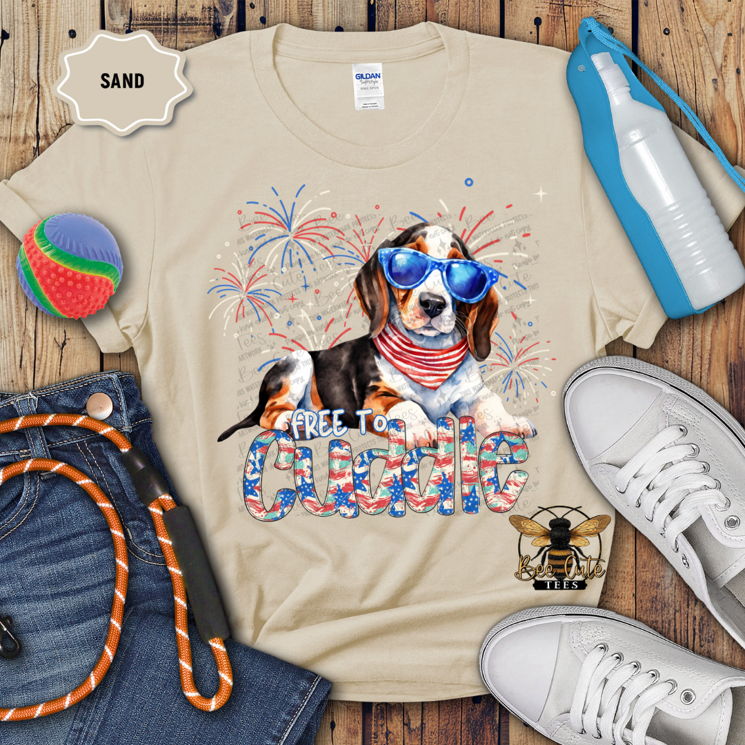 A beige colored t-shirt with a graphic print of a beagle dog wearing sunglasses and a patriotic neck collar, surrounded by Fourth of July themed decorations such as a frisbee, water bottle, and a rawhide toy. Sneakers and a crewneck sweatshirt are also displayed.
