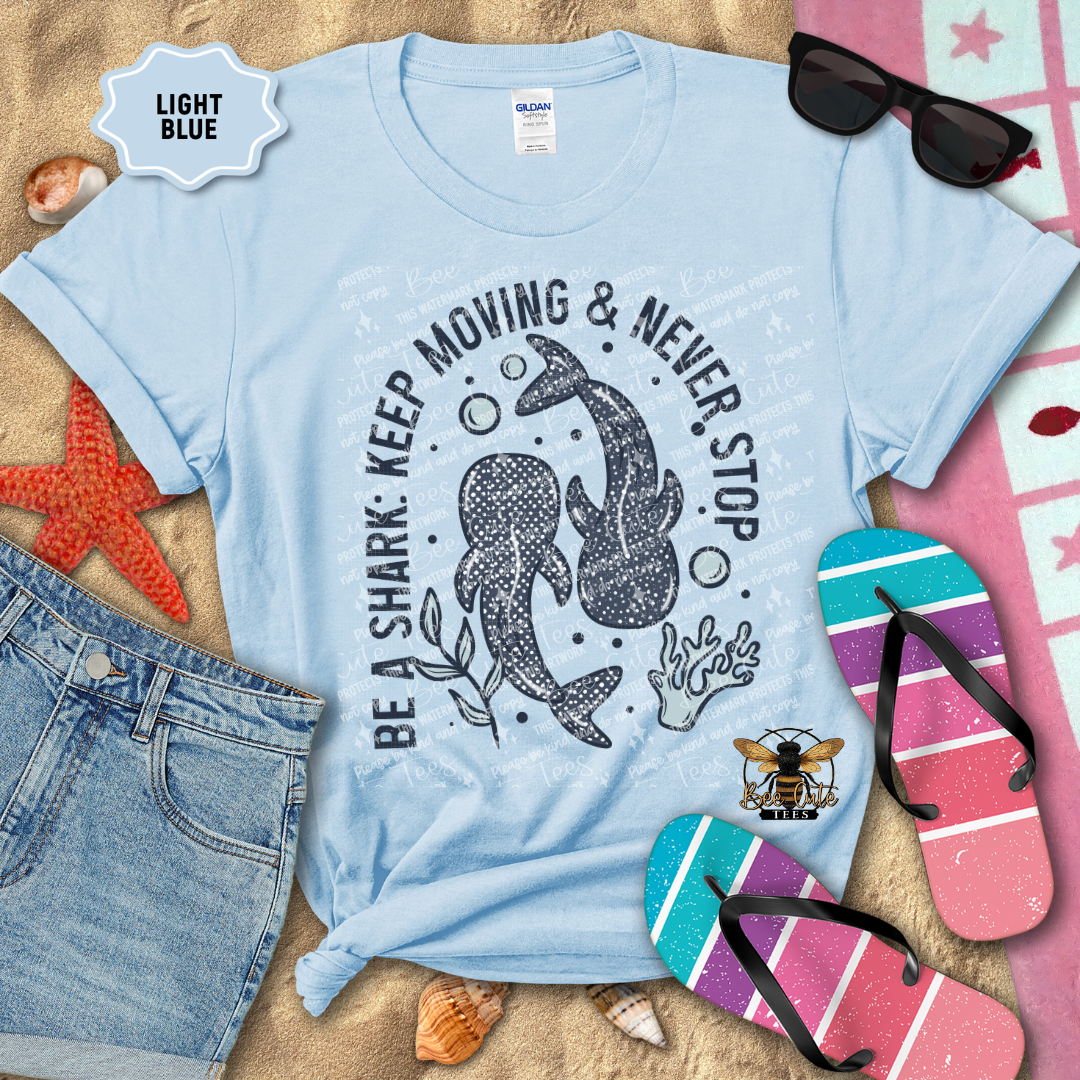 Light blue t-shirt with shark graphic and text on a sandy background with flip-flops and sunglasses.