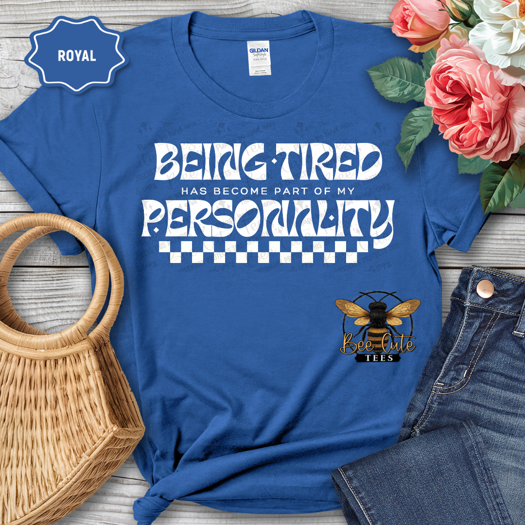 Blue t-shirt with 'Being Tired has become part of my personality' text and bee logo, placed on a wooden surface with flowers and jeans.