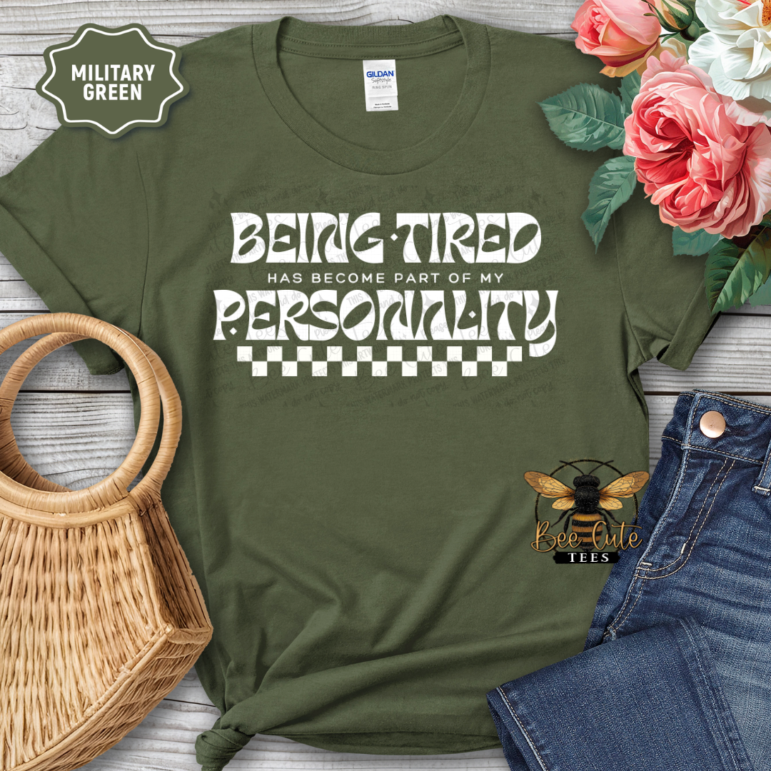 Military green t-shirt with 'Being Tired Has Become Part of My Personality' text, surrounded by flowers and casual items.