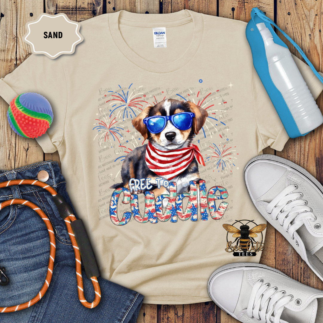 A beige colored t-shirt with a graphic print of a Bernese Mountain Dog wearing sunglasses and a neck tie, surrounded by Fourth of July themed decorations, placed on a wooden surface accompanied by a pair of white sneakers and a blue water bottle.