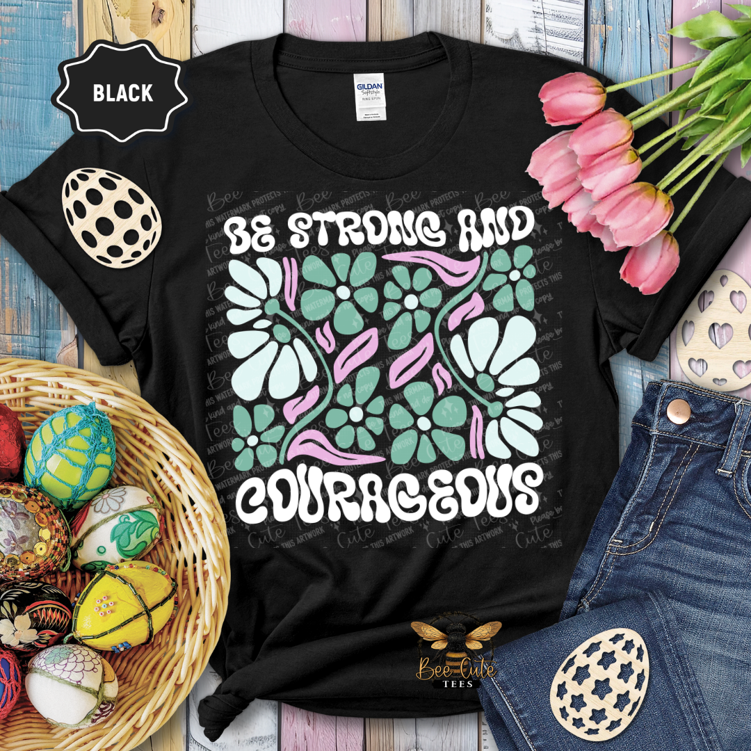 Black t-shirt with floral design and text 'Be Strong and Courageous' on a wooden surface with flowers and eggs.