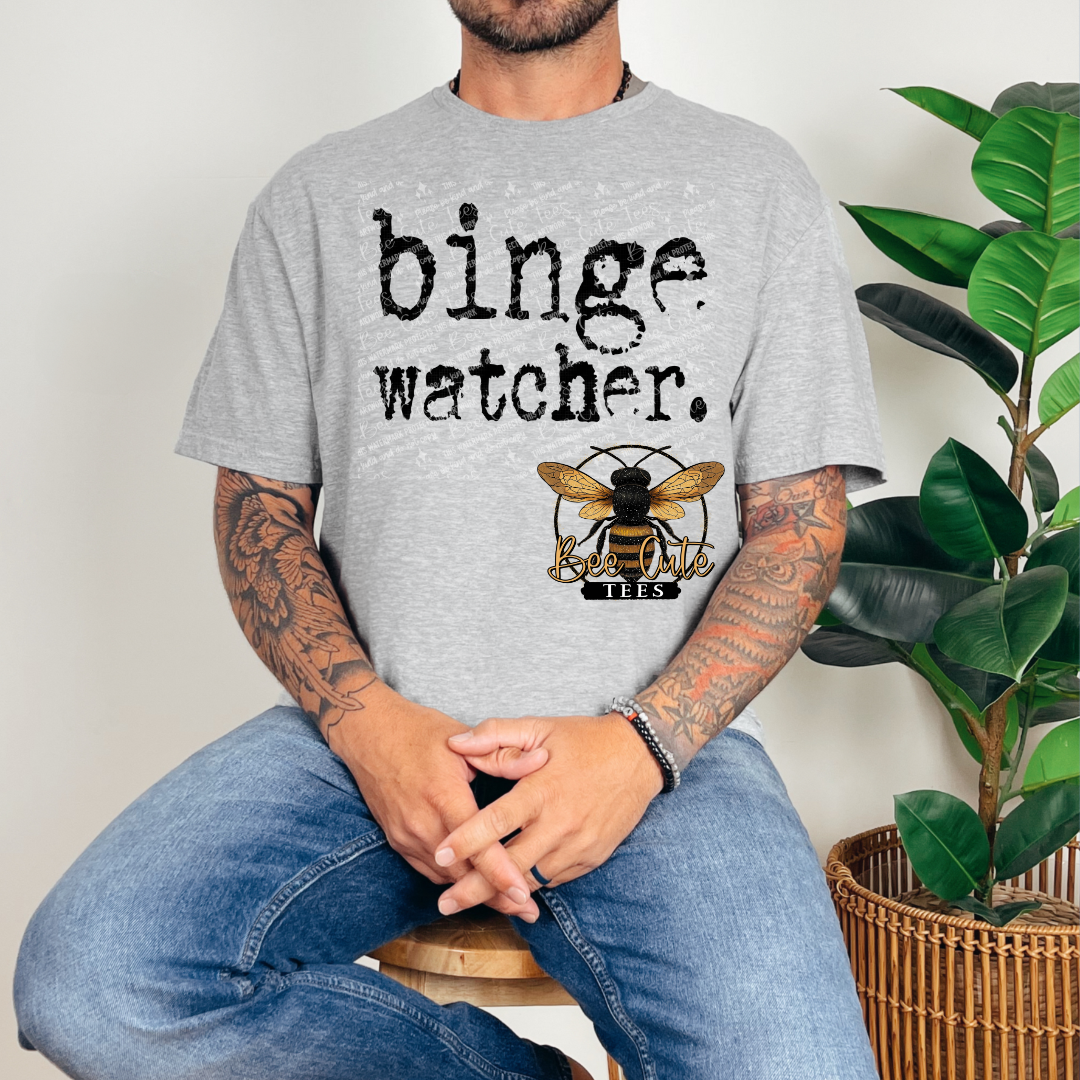 Person wearing a gray t-shirt with 'binge watcher' text and a bee logo, sitting next to a plant.