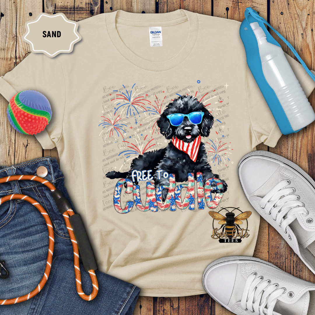 A beige colored t-shirt with a graphic print of a black doodle dog wearing sunglasses and a text 'Free To Cuddle', surrounded by Fourth of July-themed elements like fireworks. The shirt is displayed with a pair of jeans, a pair of white sneakers, a water bottle, and a beach ball.