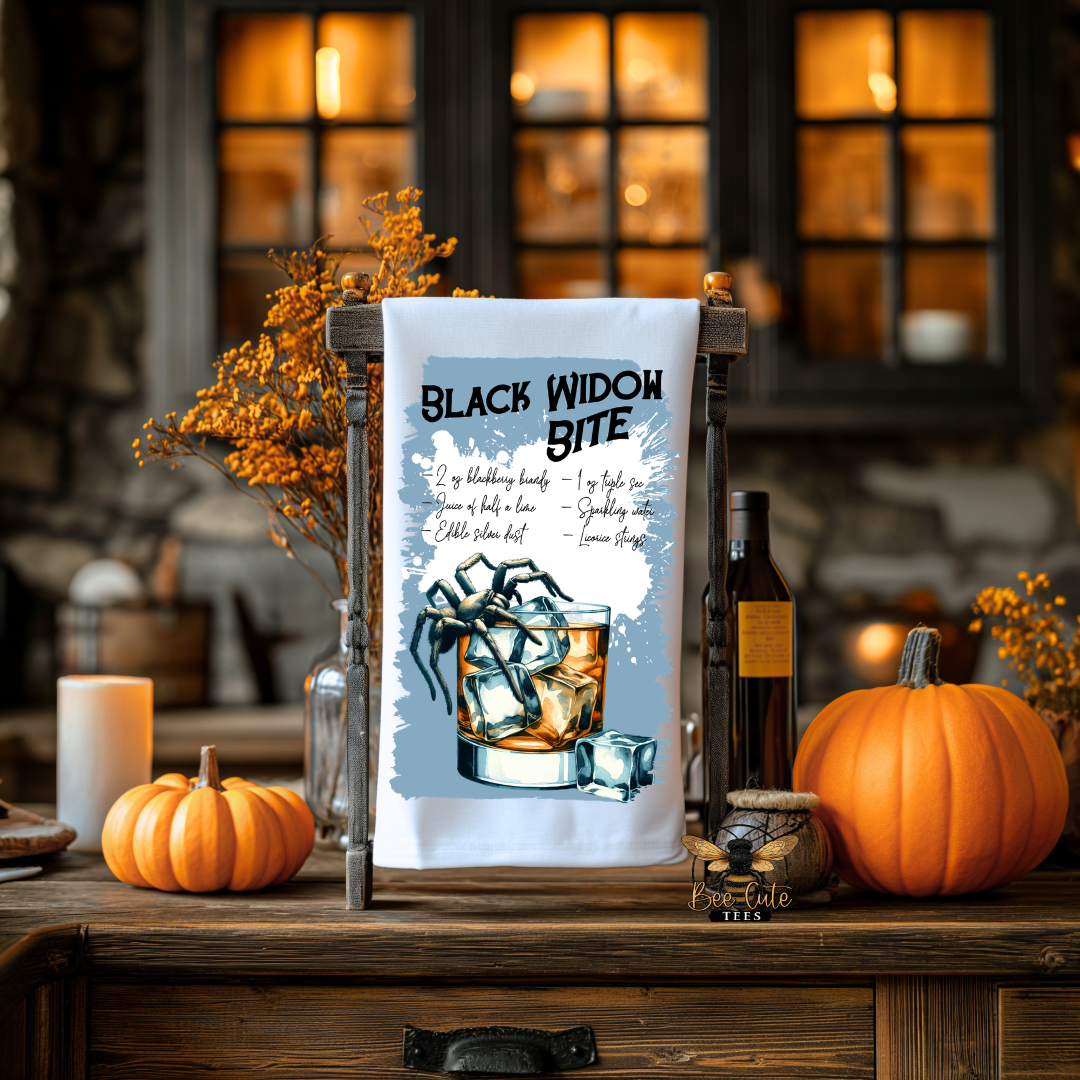 A white tea towel with a humorous 'Black Widow Bite' cocktail graphic is displayed on a rustic wooden stand. The scene is styled as a cozy autumn kitchen or bar, featuring pumpkins, a candle, a bottle, and a blurred background with warm window light.