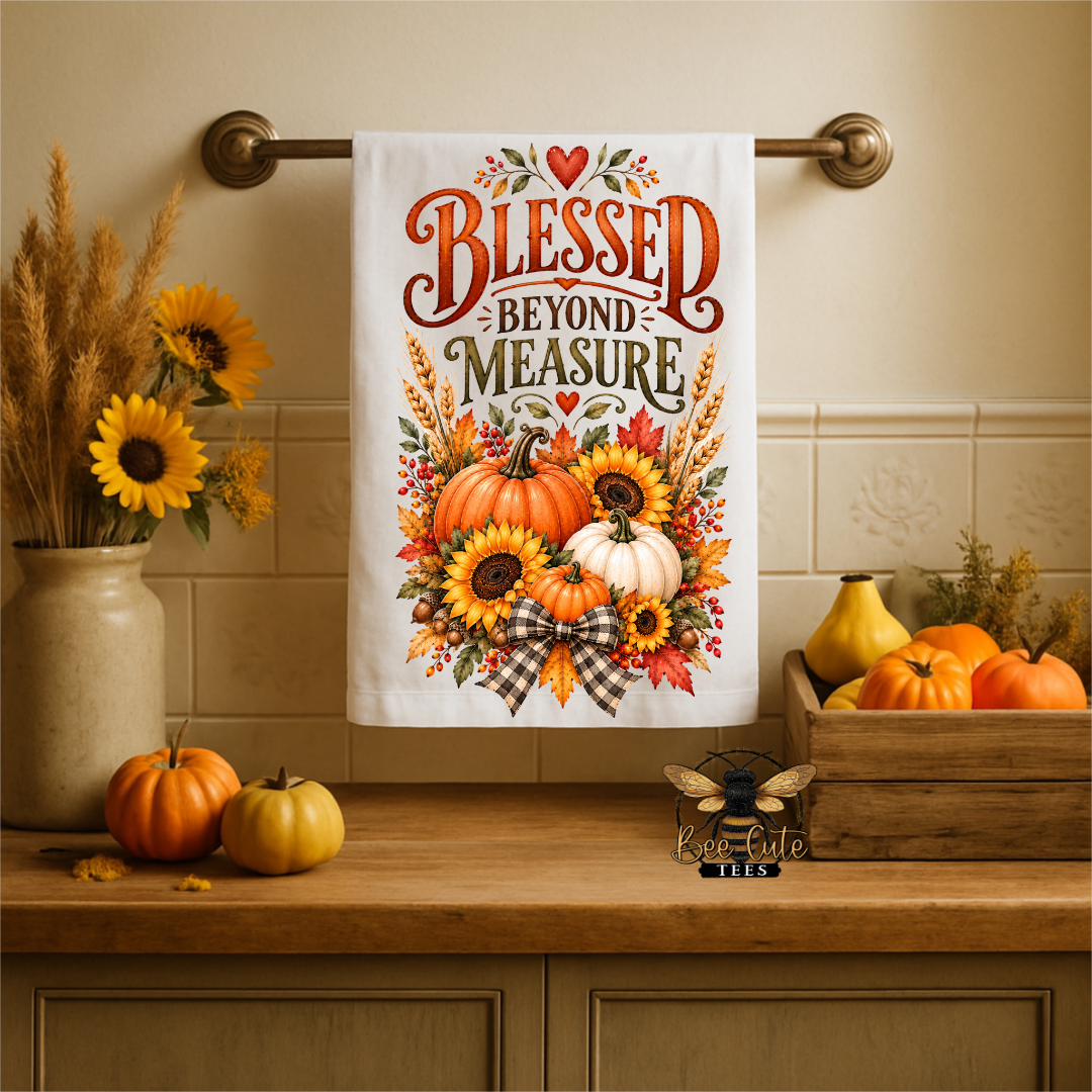 A white kitchen towel with a fall harvest design hangs on a brass towel bar. The towel features the text 'BLESSED BEYOND MEASURE' in red and brown lettering above a graphic of pumpkins, sunflowers, and autumn leaves.