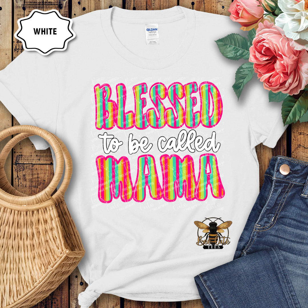 White t-shirt with colorful 'Blessed to be called MAMA' text, placed on a wooden surface with flowers and jeans.
