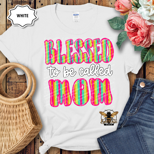 White t-shirt with colorful 'Blessed to be called MOM' text on a wooden surface with flowers and jeans.