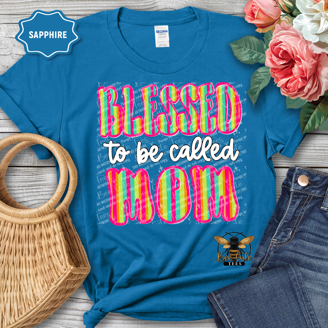 Blue t-shirt with colorful 'Blessed to be called Mom' text, surrounded by flowers and denim jeans on a wooden surface.
