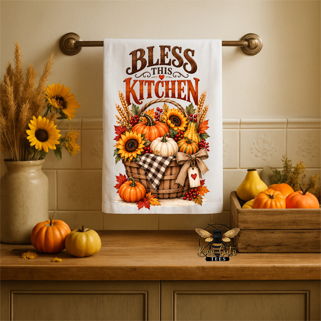 A white kitchen towel hanging on a brass rack above a wooden counter, printed with the phrase 'Bless This Kitchen' and an autumn harvest illustration featuring pumpkins, sunflowers, and fall leaves. The scene is styled with seasonal decor including pumpkins, sunflowers, and a wooden crate.