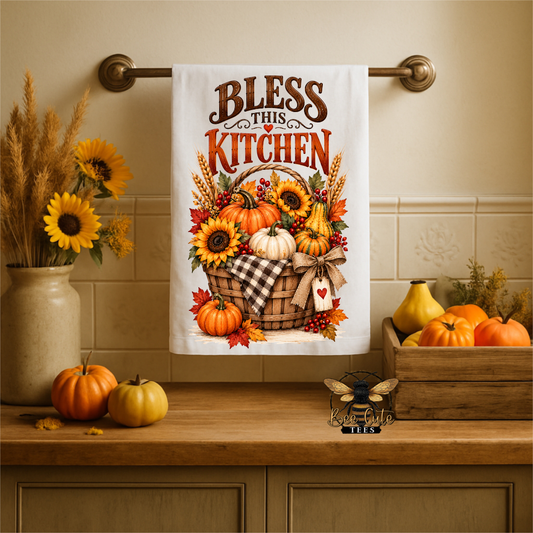 A white kitchen towel hanging on a brass rack above a wooden counter, printed with the phrase 'Bless This Kitchen' and an autumn harvest illustration featuring pumpkins, sunflowers, and fall leaves. The scene is styled with seasonal decor including pumpkins, sunflowers, and a wooden crate.