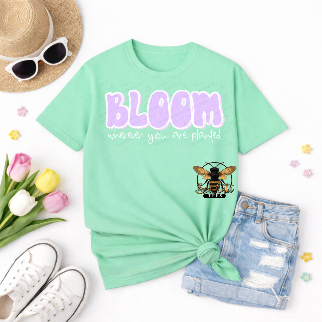 Mint green t-shirt with 'BLOOM wherever you are planted' text and bee graphic, surrounded by sunglasses, tulips, and shorts on a white background.