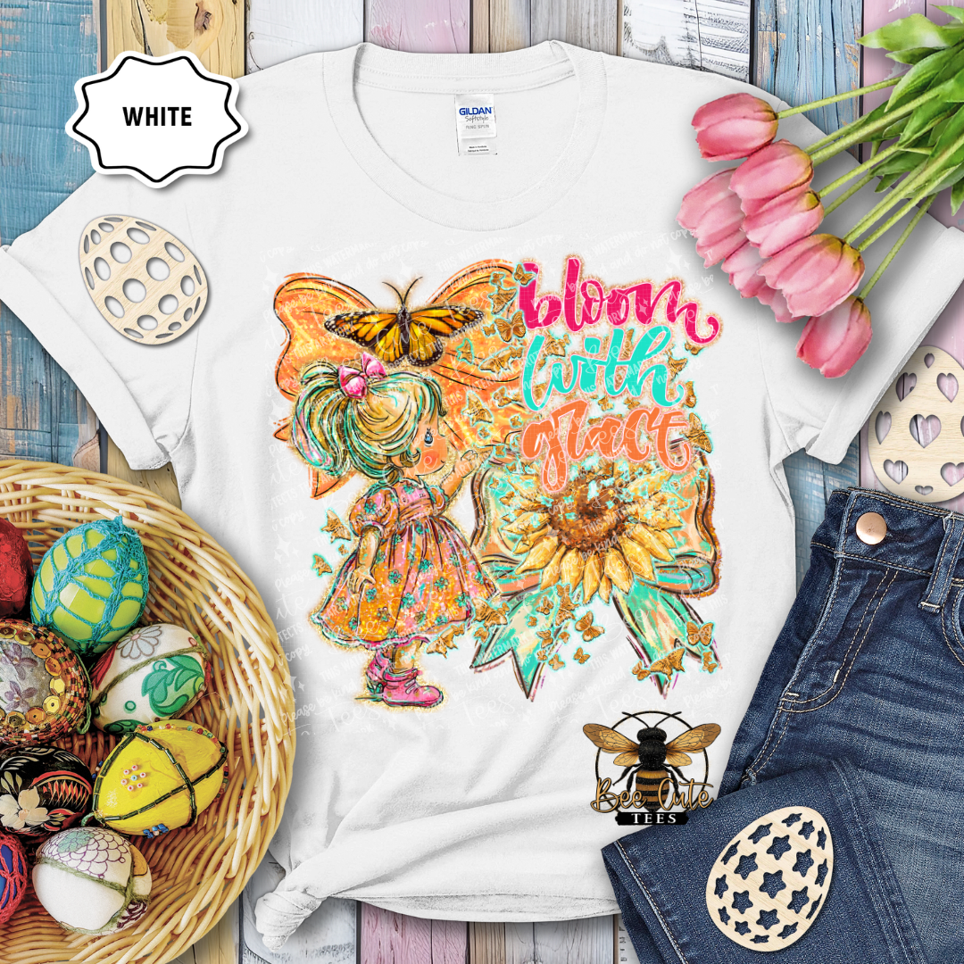 White t-shirt with a colorful design featuring a fairy, butterfly, and sunflower text 'Bloom with Flare' on a wooden surface with Easter decorations.