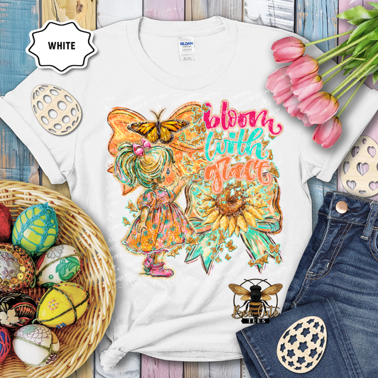 White t-shirt with a colorful design featuring a fairy, butterfly, and sunflower text 'Bloom with Flare' on a wooden surface with Easter decorations.