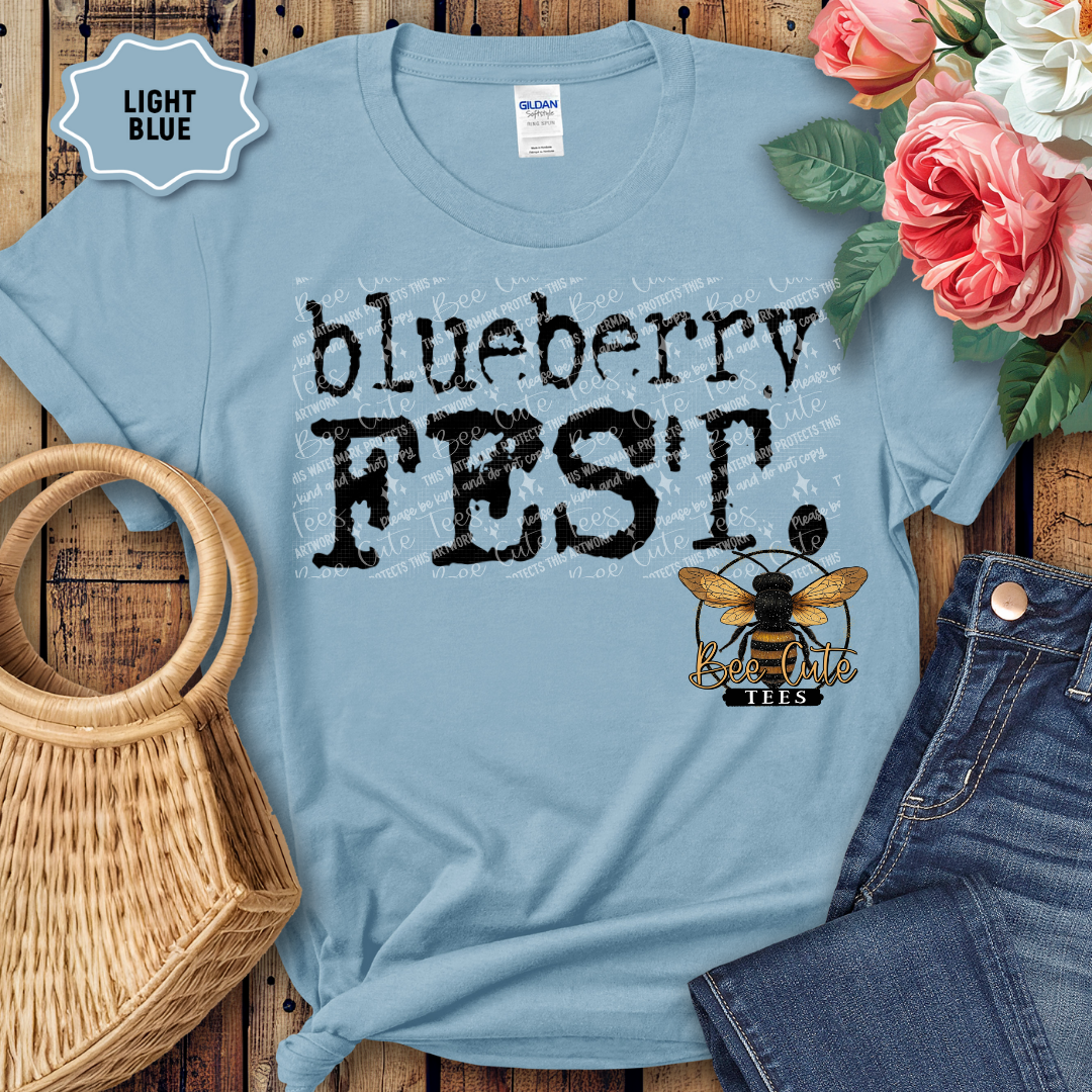 Light blue graphic t-shirt with bold black text reading “Blueberry Fest,” styled flat lay on a wooden background with denim shorts, a woven handbag, and pink flowers, featuring a Bee Cute Tees logo.
