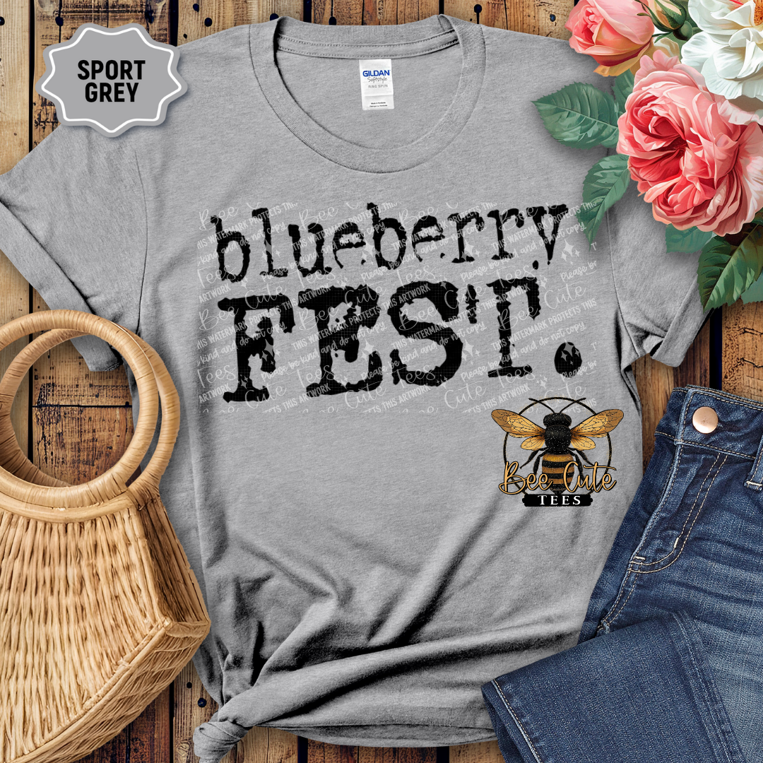 Sport Grey graphic t-shirt with bold black text reading “Blueberry Fest,” styled flat lay on a wooden background with denim shorts, a woven handbag, and pink flowers, featuring a Bee Cute Tees logo.
