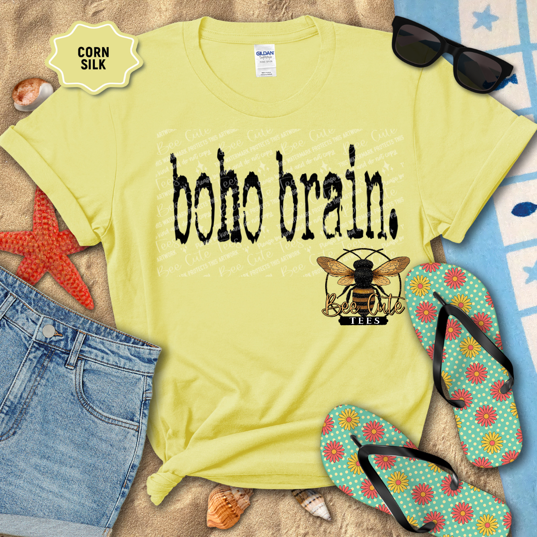 Corn silk yellow graphic t-shirt featuring distressed black “Boho Brain” text, styled flat lay on a beach towel with denim shorts, flip flops, sunglasses, seashells, and a starfish for a relaxed summer vibe.
