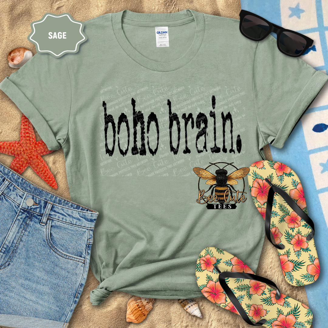 Sage green graphic t-shirt featuring distressed black “Boho Brain” text, styled flat lay on a beach towel with denim shorts, flip flops, sunglasses, seashells, and a starfish for a relaxed summer vibe.