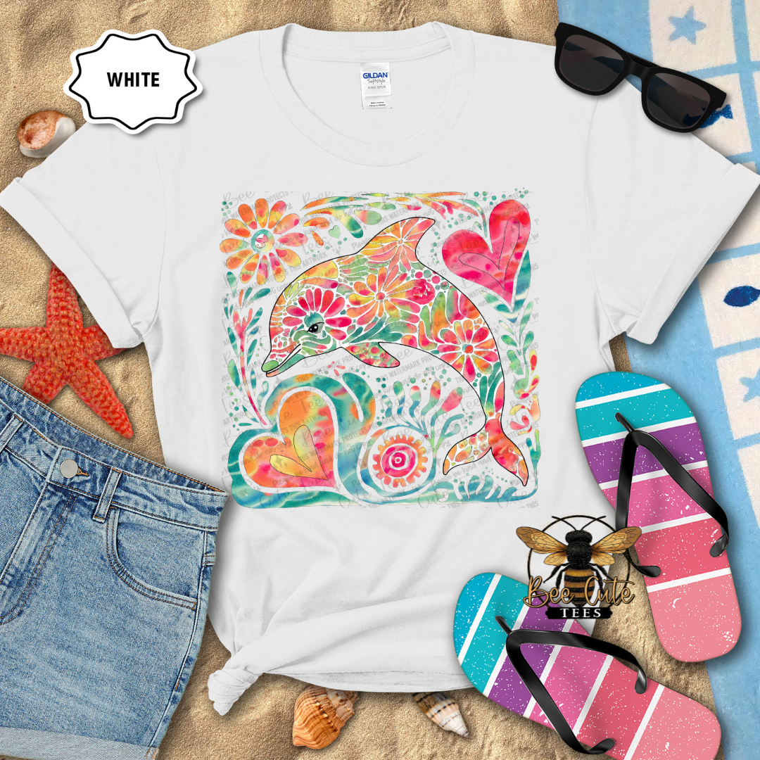 White t-shirt with a colorful dolphin design on a beach-themed background with flip-flops and sunglasses.