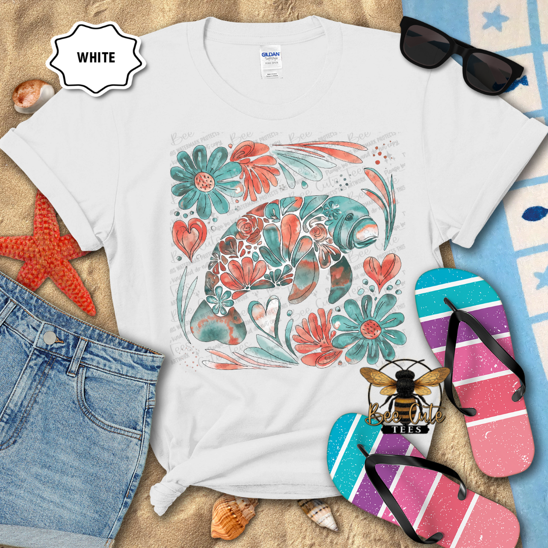 White t-shirt with floral manatee design on a beach-themed background with flip-flops and sunglasses.