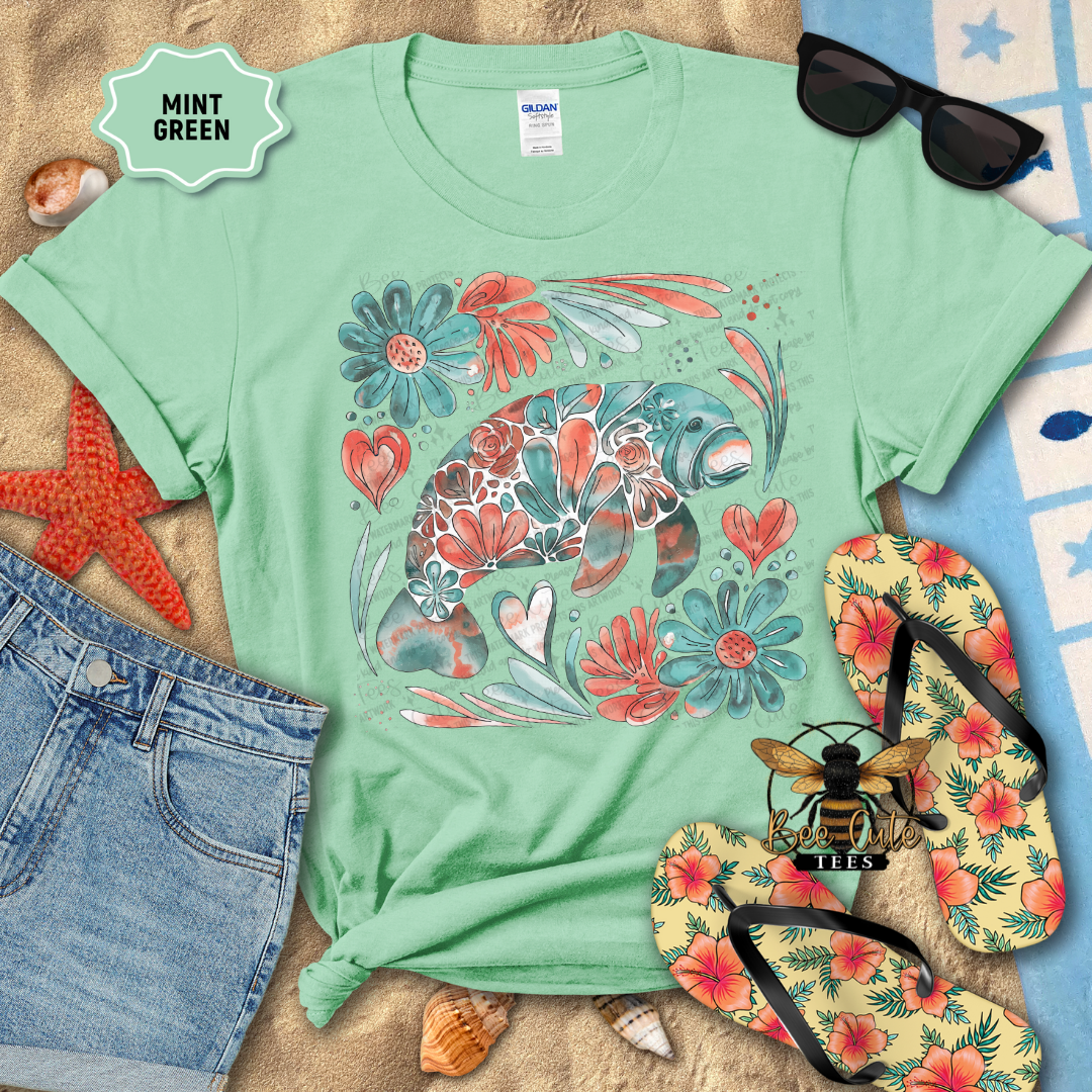 Mint green t-shirt with floral manatee design on a beach-themed background with flip-flops and sunglasses.