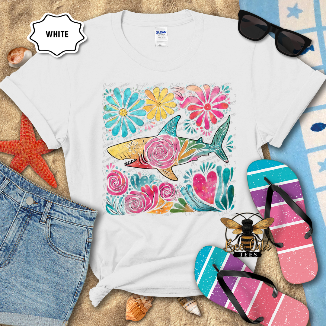 White t-shirt with colorful shark and floral design on a sandy surface with beach accessories.