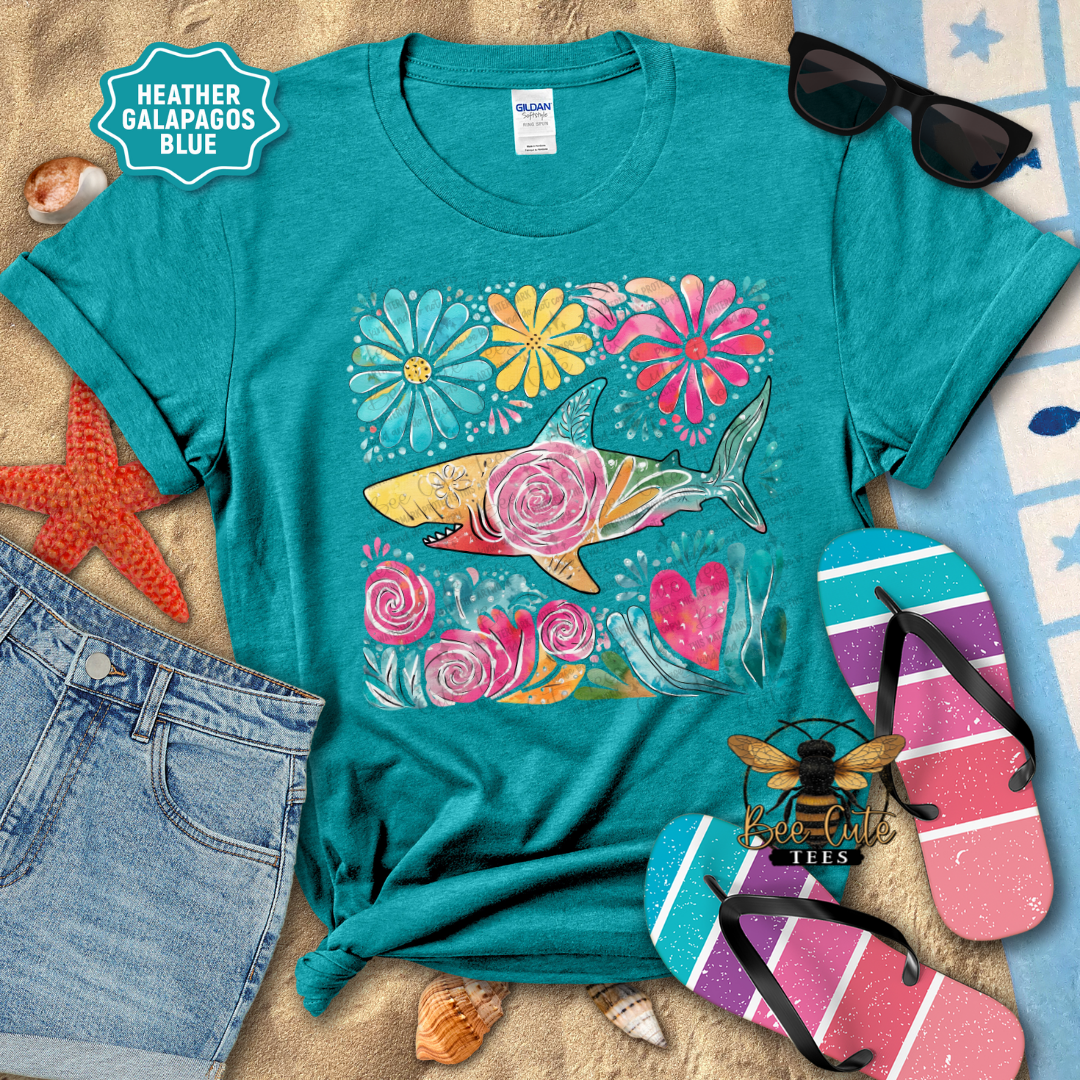 Teal t-shirt with a colorful shark and floral design on a textured surface with sunglasses and flip-flops.