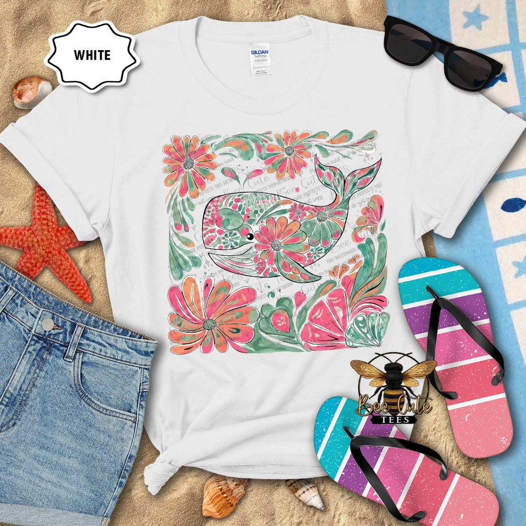 White t-shirt with a colorful whale and floral design on a sandy surface with beach accessories.