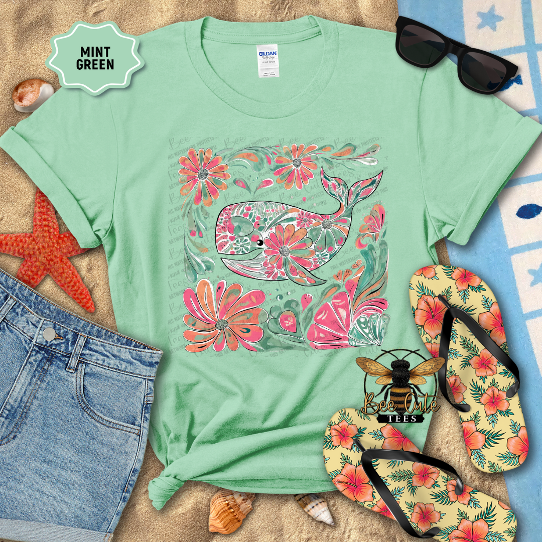 Mint green t-shirt with floral whale design on a sandy surface with beach accessories.