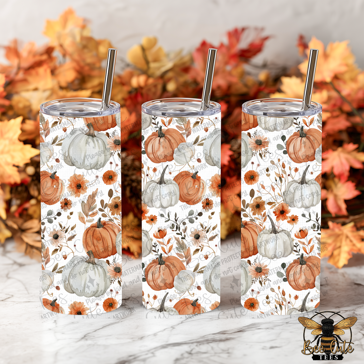 Three tall skinny tumblers with clear lids and metal straws, decorated with a white background and a repeating autumn pumpkin and floral pattern in orange, gray, and brown. The tumblers are arranged on a white marble surface with a blurred background of fall leaves.