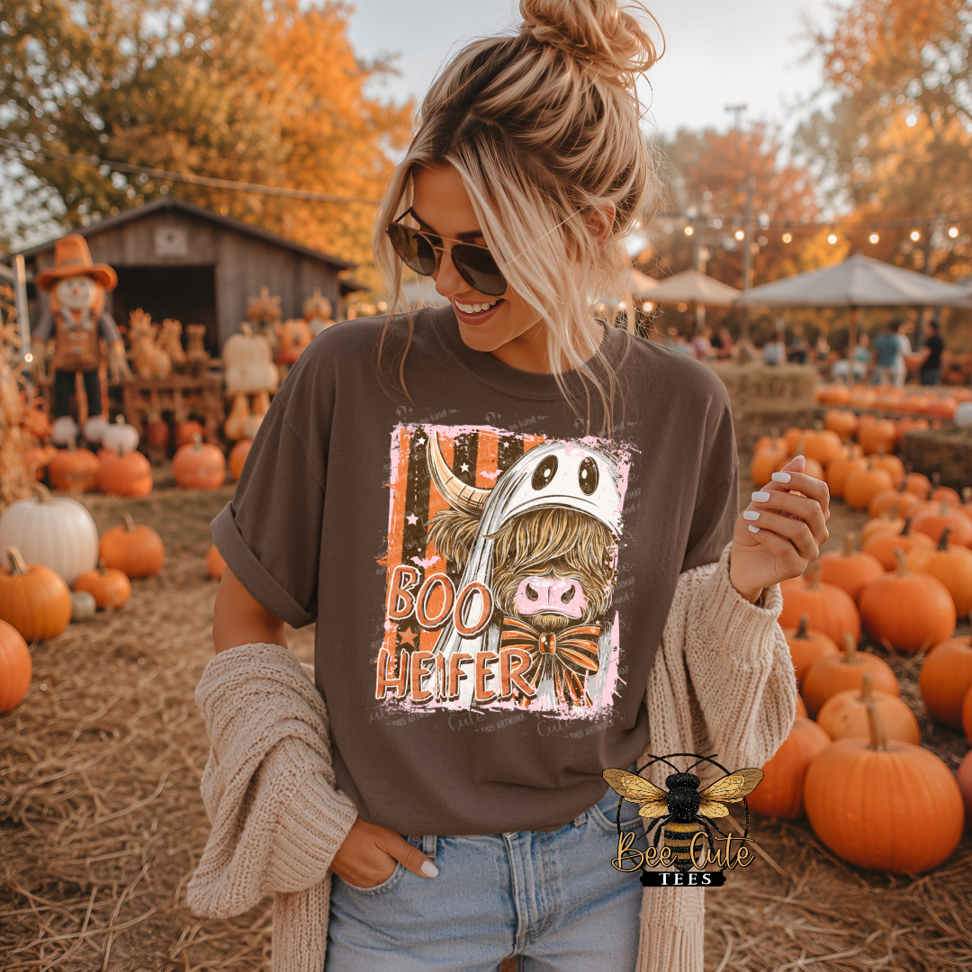 A woman wearing a brown graphic t-shirt with a 'Boo Heifer' cartoon design, styled with a beige knit cardigan and sunglasses, standing in a pumpkin patch at a fall harvest festival.