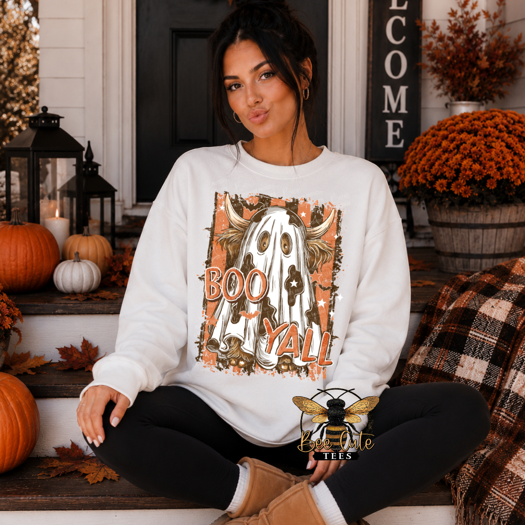 A woman sits cross-legged on a front porch wearing a white crewneck sweatshirt with a large 'Boo Y'all' Halloween graphic featuring a ghost and pumpkins. The background includes pumpkins, mums, a 'Welcome' sign, and a plaid throw blanket.