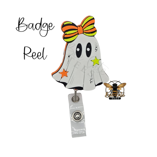 A Halloween-themed badge reel featuring a ghost design with a striped orange and yellow bow, a green star, and an orange star, attached to a silver metal clip. A small bee pin is shown next to it on a plain white background with 'Badge Reel' text on the left.