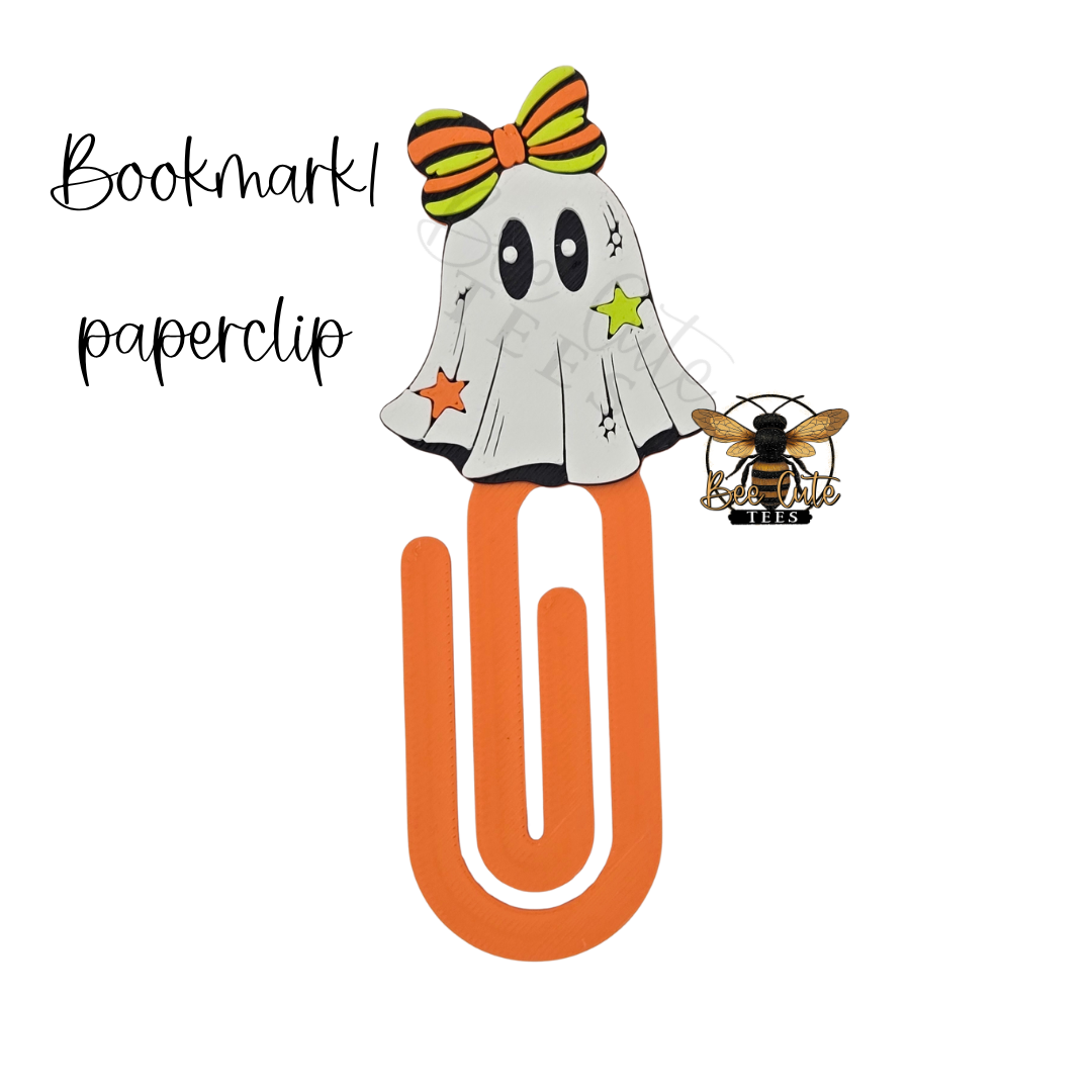 A Halloween-themed paperclip bookmark design featuring a white ghost with black eyes, a green and orange striped bow, stars, and a small bee logo, with the text 'Bookmark paperclip' on a white background.