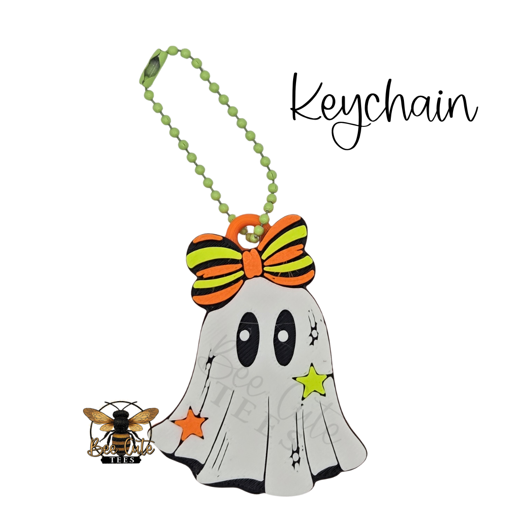 A Halloween-themed keychain featuring a cute ghost charm with a striped orange and green bow, a green beaded keychain strap, and star accents, shown on a white background with a bee logo and the word 'Keychain'.
