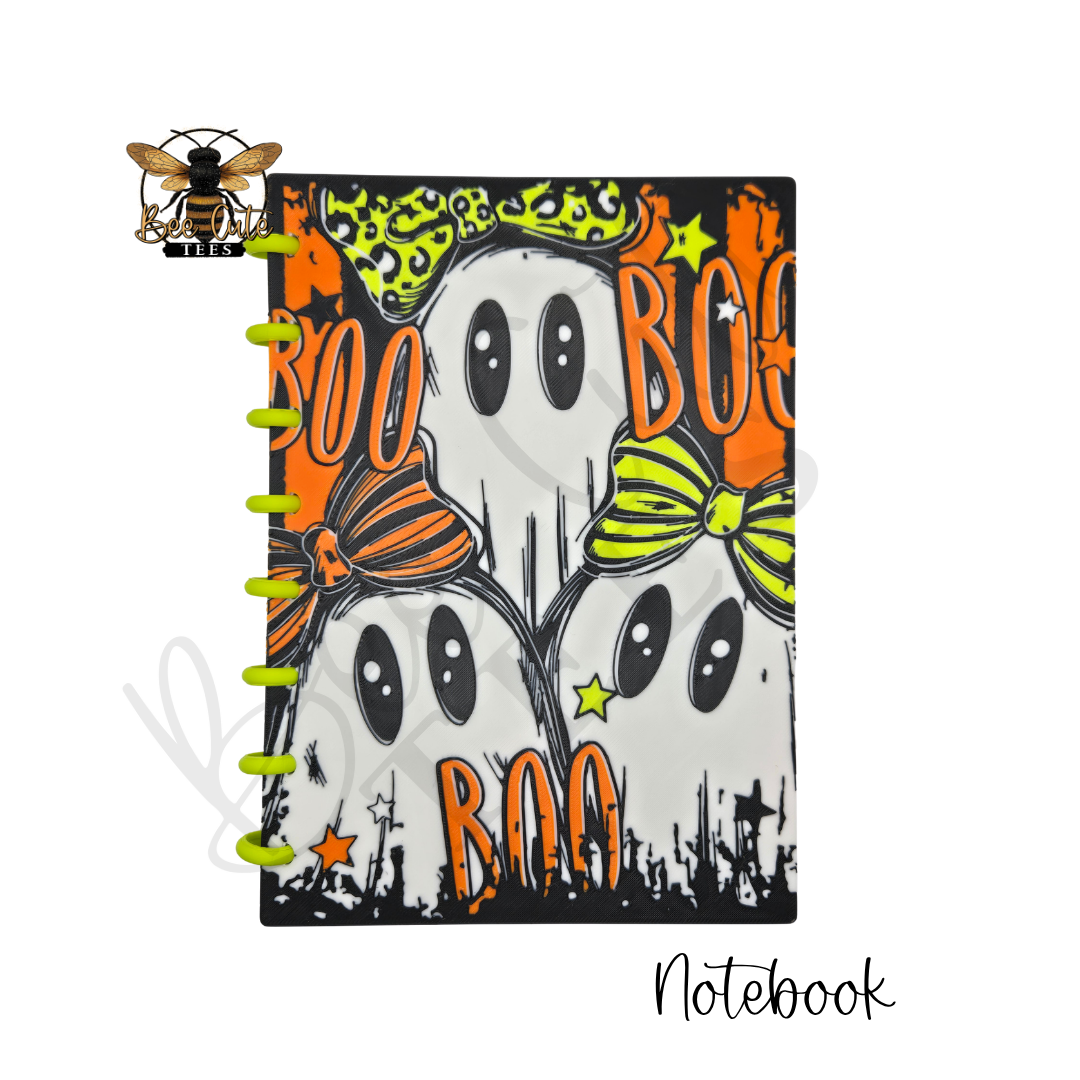 A spiral-bound notebook with a Halloween design featuring three cartoon ghosts wearing orange and green bows, with 'BOO' text and stars on a black background. A bee logo with the text 'Bee Cute Tees' appears in the top-left corner, and 'Notebook' is written in script at the bottom-right.