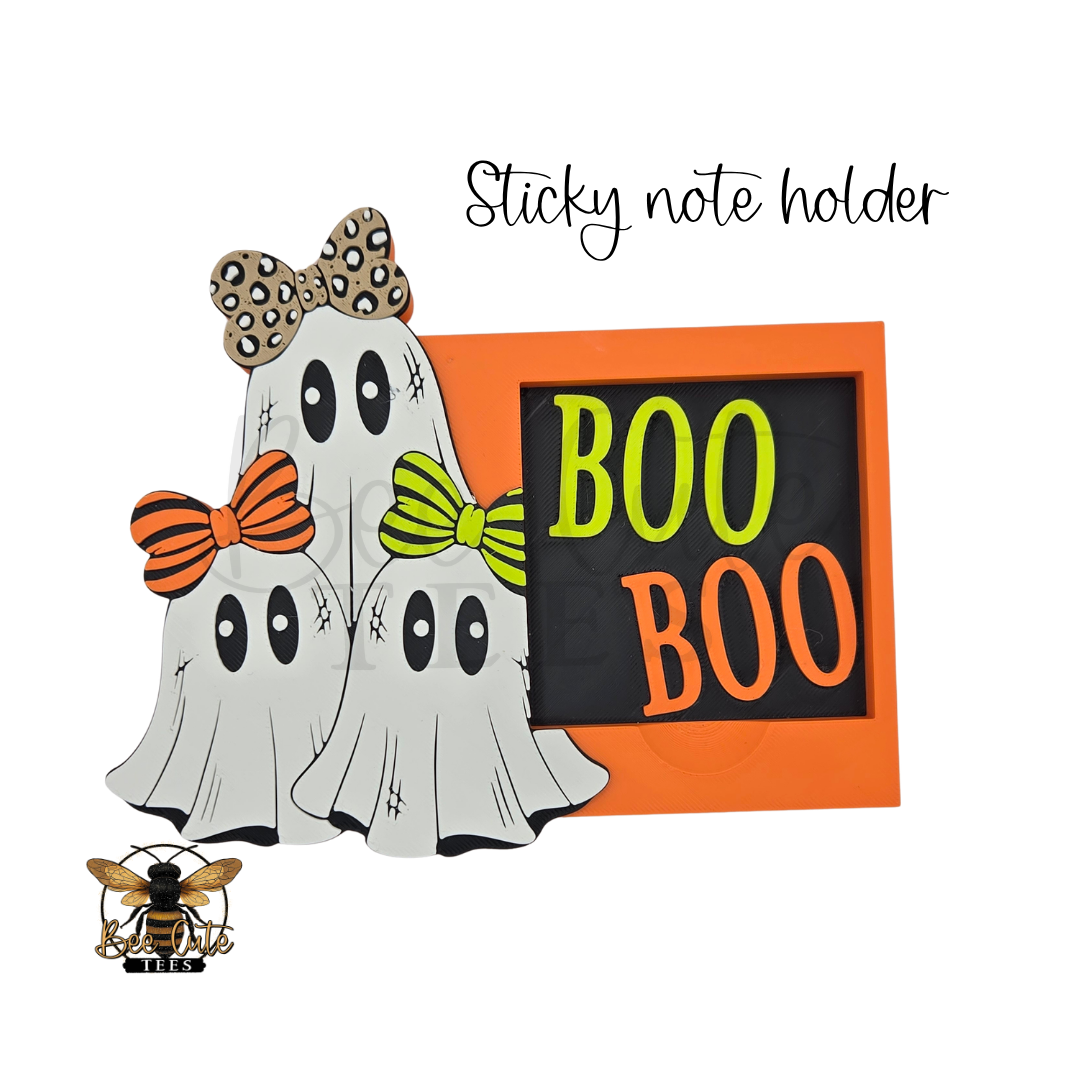 A Halloween-themed sticky note holder featuring three white ghost figures with black eyes and three colorful bows in leopard print, orange, and lime green, next to an orange frame with 'BOO BOO' written on a black background. The text 'Sticky note holder' appears above, and a bee logo is in the bottom-left corner.