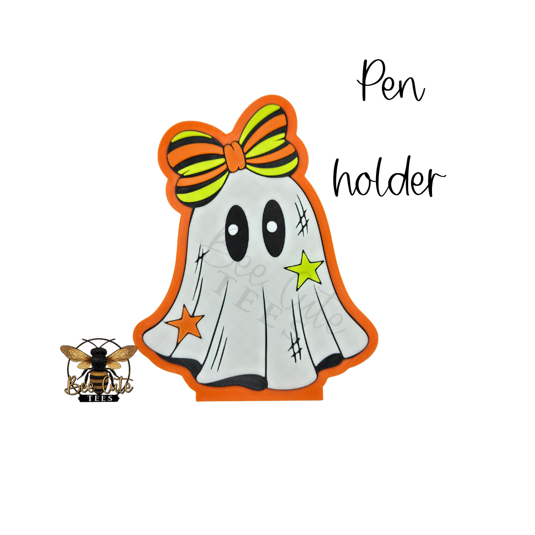 A ghost-shaped pen holder with a Halloween theme, featuring a white ghost body, orange border, black eyes, a green and orange striped bow, and star accents, shown on a white background with the text 'Pen holder' and a bee logo.